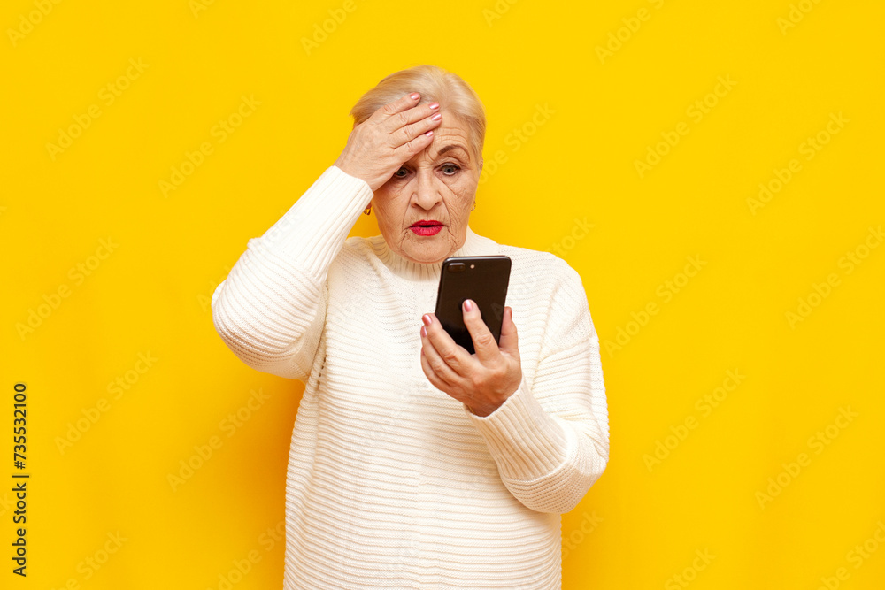 Confused old grandmother in a white sweater uses a smartphone and shows a facepalm on a yellow isolated background, sad elderly woman pensioner plays online on her mobile and worries Confused old grandmother in a white sweater uses a smartphone and shows a facepalm on a yellow isolated background, sad elderly woman pensioner plays online on her mobile and worries