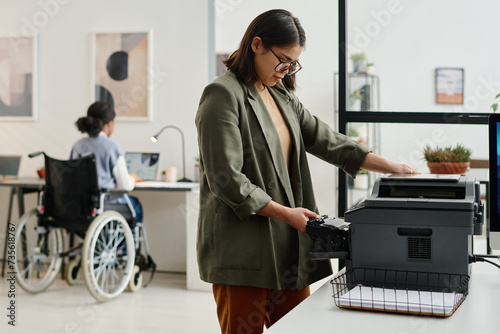 Young woman wearing smart casual clothes standing in front of printer taking out toner cartridge, her colleague in wheelchair working in background photo