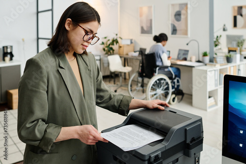 Young female manager wearing stylish eyeglasses printing documents, her colleague with disability working in background photo