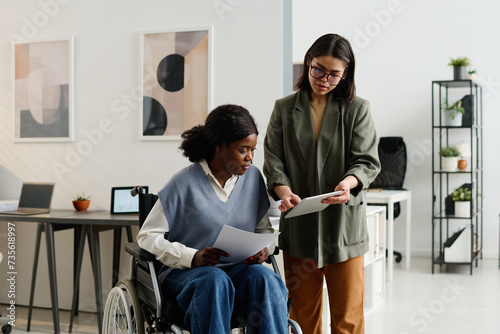 Medium long shot of young Caucasian woman showing data on digital tablet to her African American coworker with disability photo