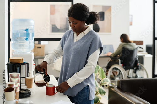 Selective focus shot of young African American woman pouring coffee into mug during break at work in modern office photo