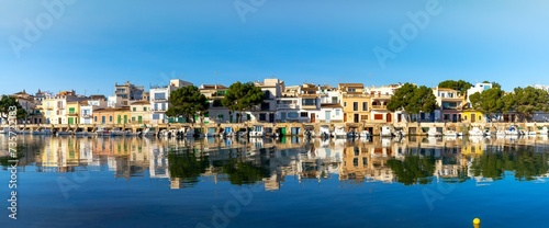 panorama cityscape view of Portocolom with colourful and whitewashed houses and boat garages on the waterfront with reflections