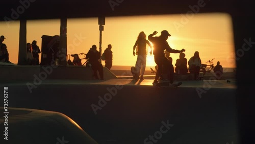 Silhouettes of skateboarders hanging out and riding skateboards in front of sunset sun at Venice Beach skate park in Los Angeles, California. Slow motion shot.