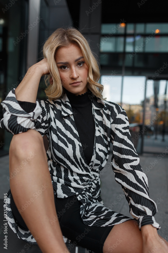 Fashion happy beautiful young girl with a smile in a fashionable zebra print dress and shorts with white sports sneakers sits and rest in the city Fashion happy beautiful young girl with a smile in a fashionable zebra print dress and shorts with white sports sneakers sits and rest in the city