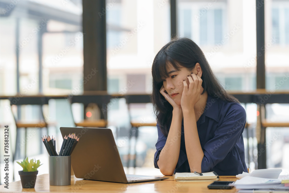 Asian woman working with a laptop while sitting at a laptop in the office, thinking of professional plans, project management, and considering new business ideas.