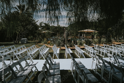 
Plano general de la decoración para una boda judía a celebrar en un jardín privado. Vemos un pasillo central con una alfombra blanca y a los lados dos filas de sillas. Entorno natural con cactus.