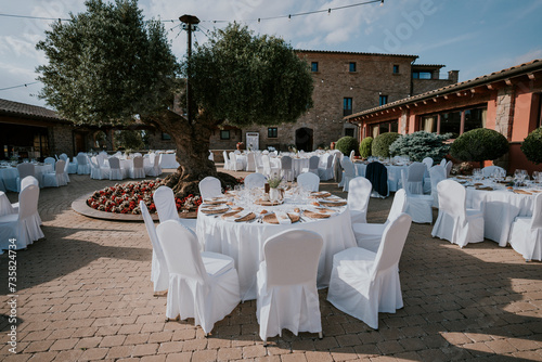 
Banquete de bodas en el exterior de la masía. Mesas decoradas con manteles blancos, sillas blancas, cristalería transparente. Entorno natural, olivo, piedra y adoquines. Día de verano. Plano general. photo