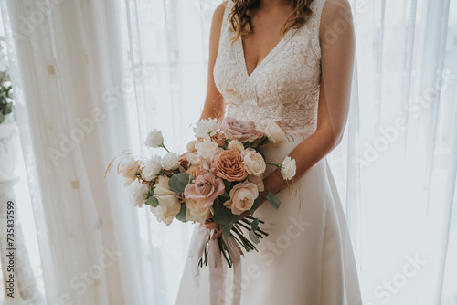 
La joven novia sostiene un hermoso ramo de rosas blancas y rosa pálido. La hermosa novia está al lado de una gran ventanal. Fotografía horizontal con tonos blancos. photo
