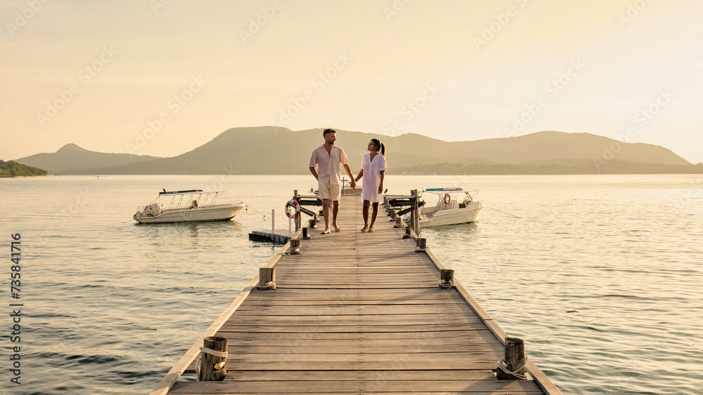 couple walking on a wooden pier in the ocean at sunset in Thailand.Caucasian men and Asian woman diverse couple waking at a jetty in the ocean, man and women watching sunset together couple walking on a wooden pier in the ocean at sunset in Thailand.Caucasian men and Asian woman diverse couple waking at a jetty in the ocean, man and women watching sunset together