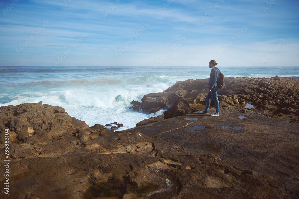A man traveler with a backpack admiring the seascape scenery from a rocky cliff, watching waves crushing on the headland