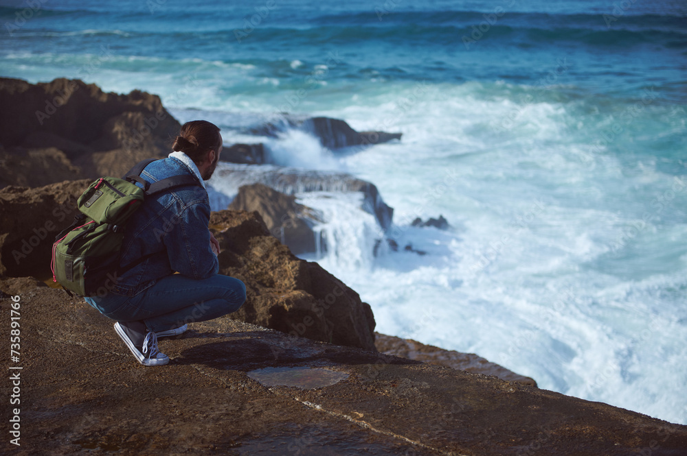 Absorbed male traveler with backpack, admiring the dangerous splashing waves from a rocky cliff