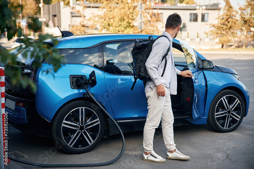 With backpack. Man with blue electric car on the charge station © standret