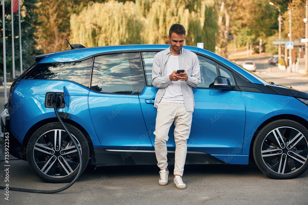 Blue vehicle, holding smartphone. Man with blue electric car on the charge station Blue vehicle, holding smartphone. Man with blue electric car on the charge station