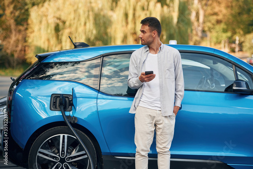 Blue vehicle, holding smartphone. Man with blue electric car on the charge station © standret