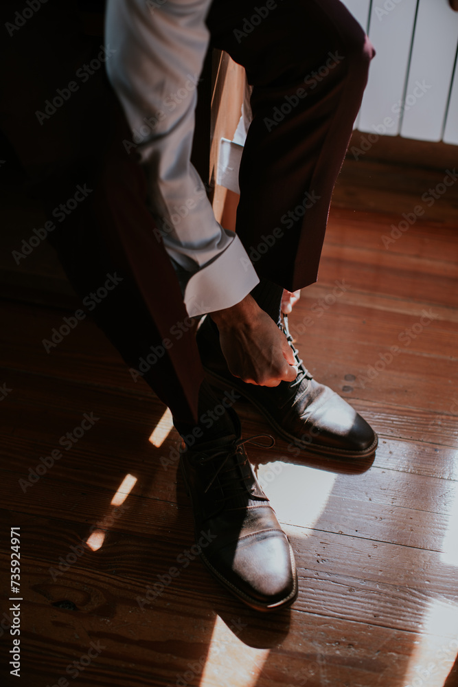 Hombre atando los cordones de sus zapatos. Está sentado en una silla y entra un rayo de luz. Detalle plano de zapatos de cuero. Foto vertical en tonos marrones.
