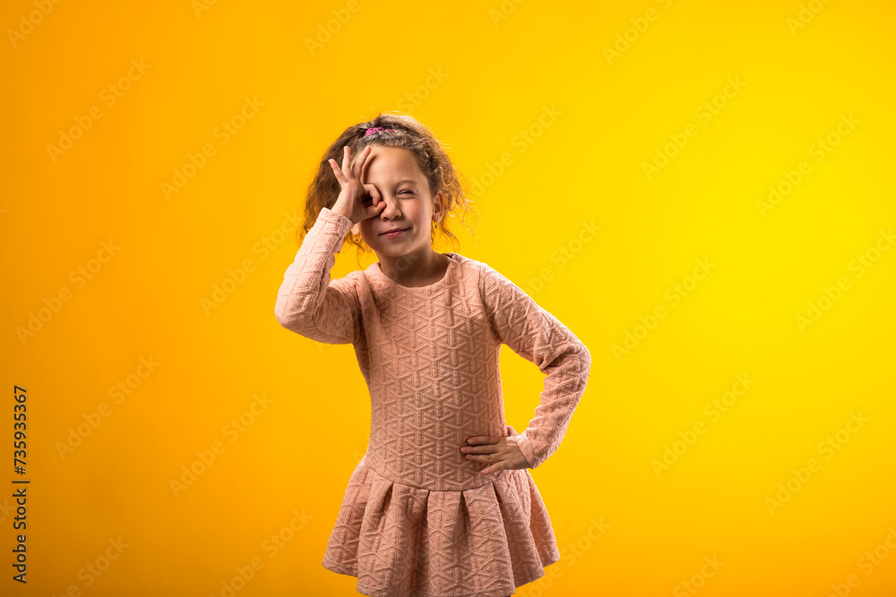 Smiling child girl looking through ok gesture over yellow background. Positive emotions concept Smiling child girl looking through ok gesture over yellow background. Positive emotions concept