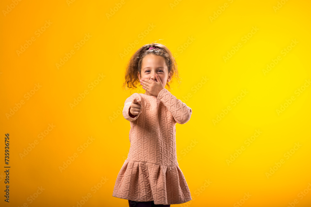 Kid girl mocking and teasing at someone showing finger at camera and closing her mouth over yellow background. Bulling concept Kid girl mocking and teasing at someone showing finger at camera and closing her mouth over yellow background. Bulling concept