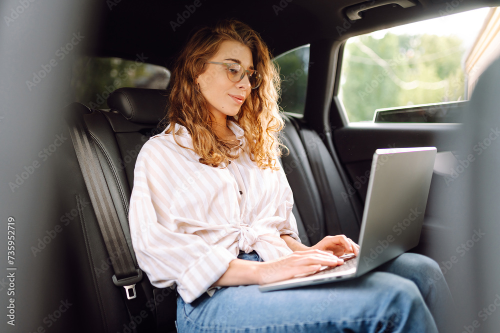 Young woman with laptop while sitting in the back seat of a car. Education online. Remote work. Business, blogging, freelance concept. Young woman with laptop while sitting in the back seat of a car. Education online. Remote work. Business, blogging, freelance concept.
