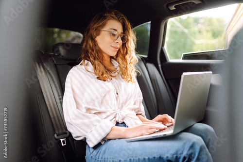 Young woman with laptop while sitting in the back seat of a car. Education online. Remote work. Business, blogging, freelance concept. © maxbelchenko
