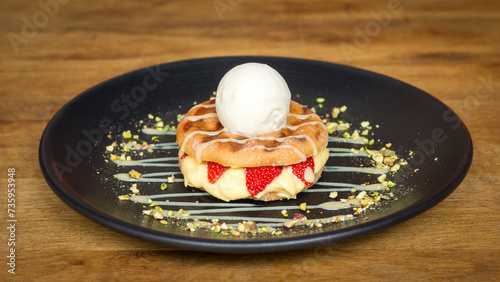 Donut dessert with strawberries, ice cream and pistachio nuts on black plate on wooden table in a restaurant