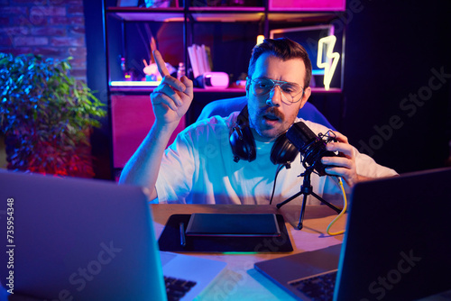 Young man  broadcaster in casual T-shirt expressing ideas into microphone in neon-lit room at home-studio. Concept of youth people with social media and smart working  lifestyle  online streaming.