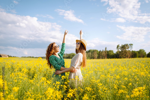 Smiling female tourists walking through flowering field, touching yellow flowers. Nature, vacation, relax and lifestyle. Summer landscape.