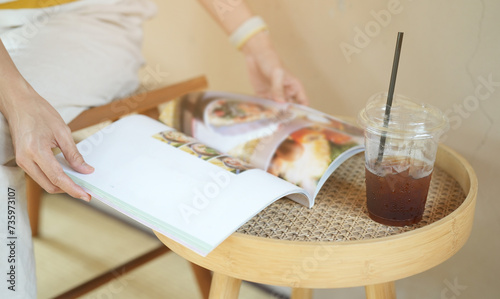 Close up hand of young girl holding and reading magazine with glass of ice coffee on wooden table in cafe