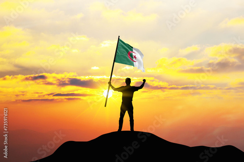 Algeria flag being waved by a man celebrating success at the top of a mountain against sunset or sunrise. Algeria flag for Independence Day.