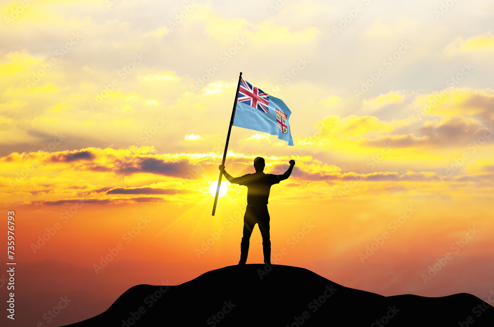 Fiji flag being waved by a man celebrating success at the top of a mountain against sunset or sunrise. Fiji flag for Independence Day.