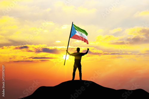 Equatorial Guinea flag being waved by a man celebrating success at the top of a mountain against sunset or sunrise. Equatorial Guinea flag for Independence Day.