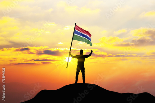 Gambia flag being waved by a man celebrating success at the top of a mountain against sunset or sunrise. Gambia flag for Independence Day.