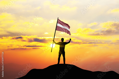 Latvia flag being waved by a man celebrating success at the top of a mountain against sunset or sunrise. Latvia flag for Independence Day. ©  minionionniloy