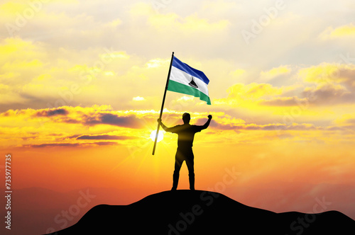 Lesotho flag being waved by a man celebrating success at the top of a mountain against sunset or sunrise. Lesotho flag for Independence Day. ©  minionionniloy