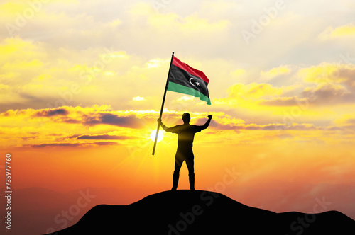 Libya flag being waved by a man celebrating success at the top of a mountain against sunset or sunrise. Libya flag for Independence Day.