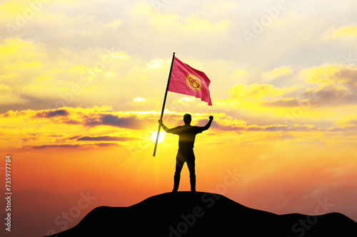 Kyrgyzstan flag being waved by a man celebrating success at the top of a mountain against sunset or sunrise. Kyrgyzstan flag for Independence Day.