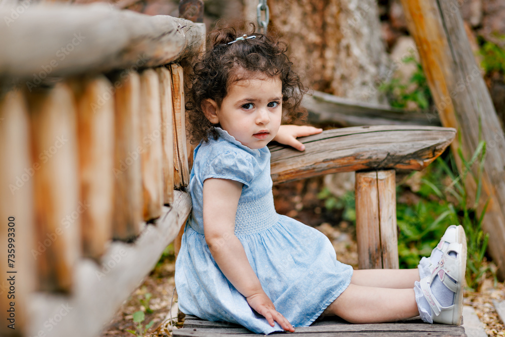 Capturing innocence. Curly-haired two-year-old girl sitting on a wooden bench in the park and looking at camera