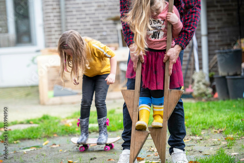 Father helping his daughter balance on stilts in their garden having fun. Her sister in the background trying to skateboard photo