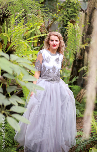 A beautiful blonde in an elegant tulle dress in a botanical garden. Background greenery and tropical plants. Beauty and fashion concept