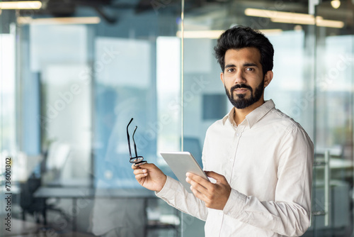 Portrait of a young Indian male programmer, developer, specialist standing in the office, holding glasses and a tablet, confidently looking at the camera