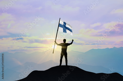 Finland flag being waved by a man celebrating success at the top of a mountain against sunset or sunrise. Finland flag for Independence Day.