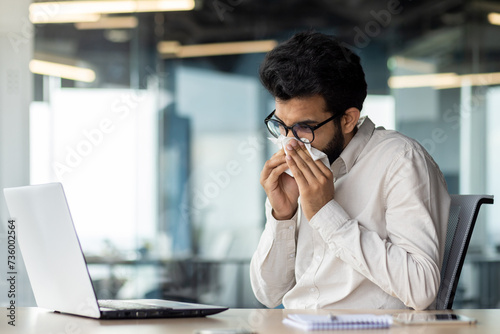 A sick young Indian man is not feeling well at the workplace. He sits at the desk in the office and wipes his nose with a napkin from a runny nose