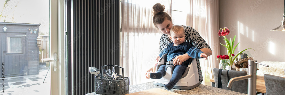 Mother feeding her small baby his lunch while he sits in his high chair on top of the kitchen side Mother feeding her small baby his lunch while he sits in his high chair on top of the kitchen side