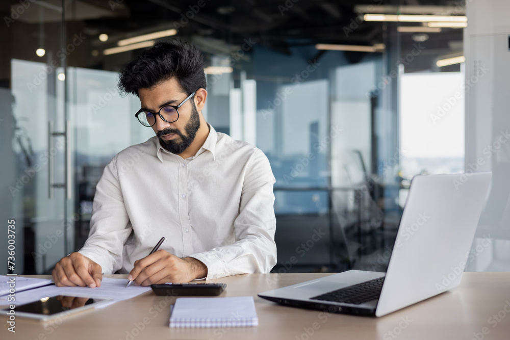 A focused and serious young Indian man works as an accountant, auditor, and financial analyst. Sitting at the desk in the office and working with accounts and data