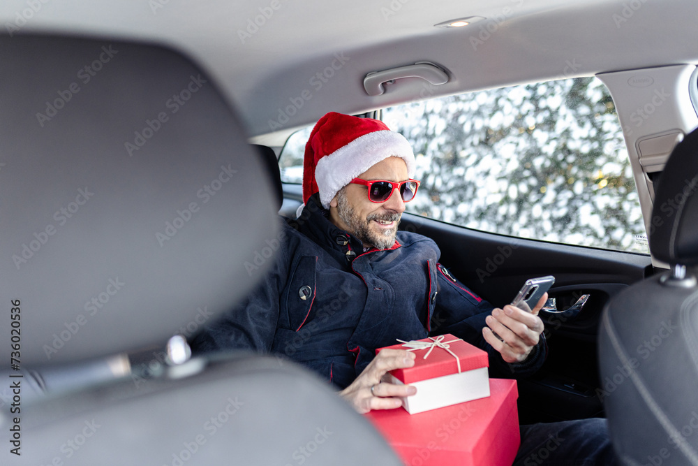 Man wearing santa claus hat and sunglasses using mobile phone and holding holiday presents while sitting on back seat in the car.