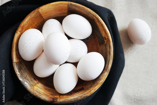 White chicken eggs in a wooden bowl on a dark towel on a light background