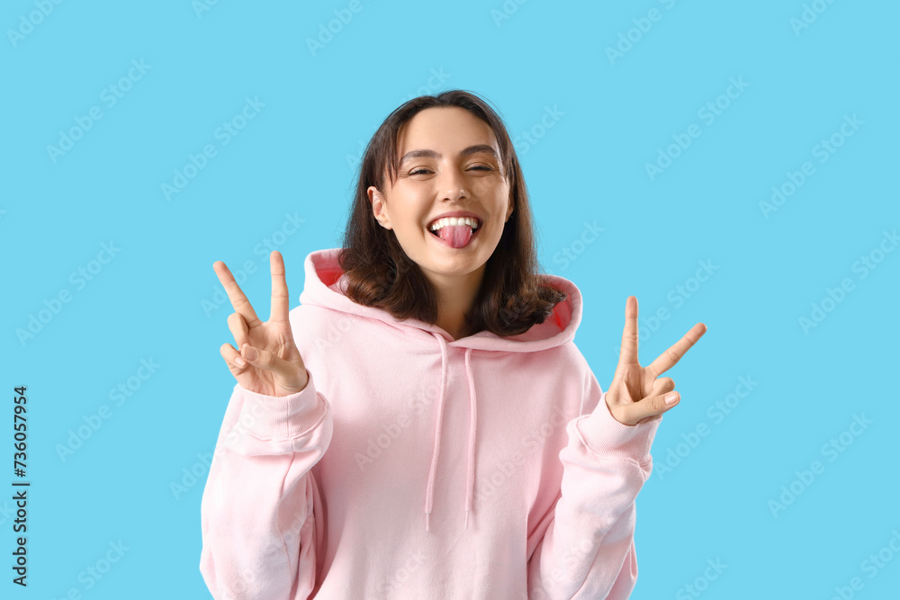 Young woman in pink hoodie showing victory gesture on blue background Young woman in pink hoodie showing victory gesture on blue background