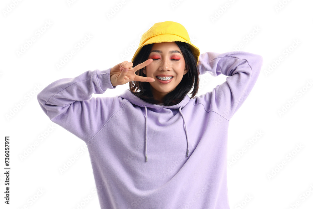 Young Asian woman in lilac hoodie and bucket hat showing victory gesture on white background Young Asian woman in lilac hoodie and bucket hat showing victory gesture on white background