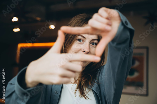 A stylish young woman with blue eyes and freckles on her face, and with a nose piercing and a blue shirt makes a frame of her fingers indoors.