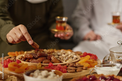 Hand of young Muslim woman taking dried date fruit from plate with variety of snacks while having festive dinner with her husband photo