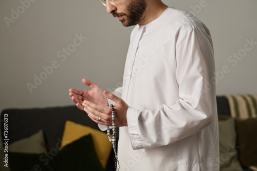 Cropped shot of young Muslim man in white kandora holding rosary beads and keeping palms open during silent pray on religious occasion photo